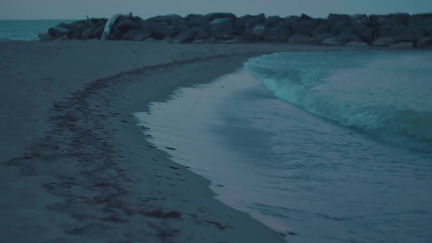 Gentle waves roll onto a sandy beach at dusk with a rocky breakwater stretching into the calm sea under a soft fading sky