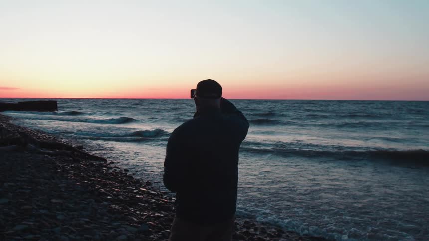 A man walks on a rocky shore at sunset taking a photo of the ocean horizon