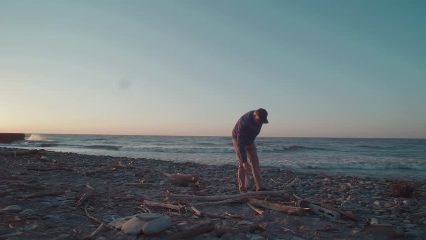 Man in cap walking on beach and picking up wooden branches