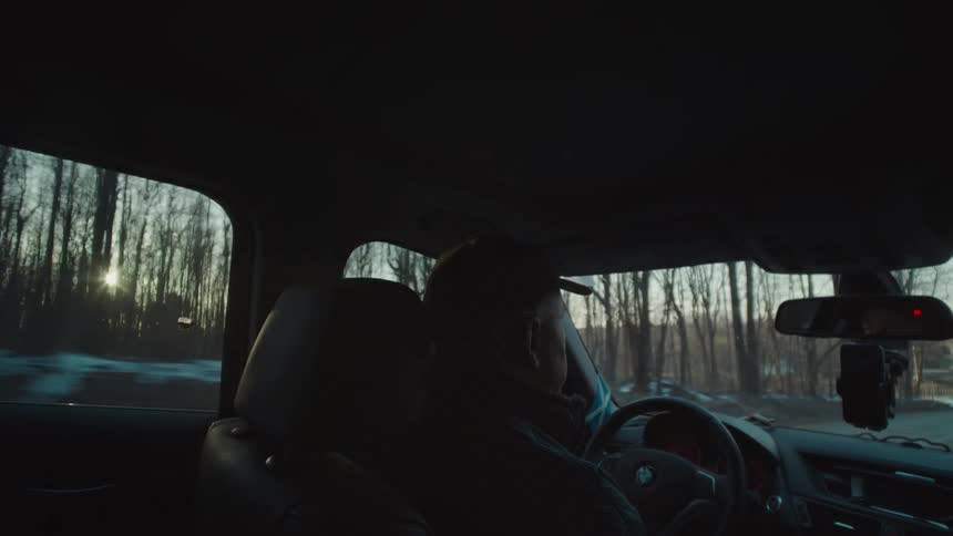 A man wearing a hat drives on a road with trees and snow on both sides around midday