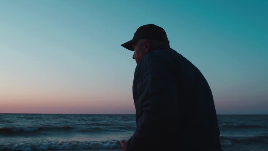 A man in a cap stands by the ocean looking out at the waves during sunset or sunrise