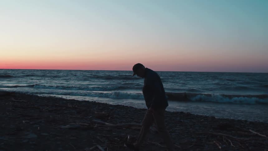 A man wearing a hat walks on the beach at sundown as ocean waves hit the shore