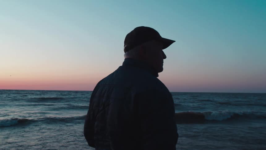 A person wearing a cap stands on a rocky beach looking out at the ocean during a colorful sunset with waves gently crashing on the shore