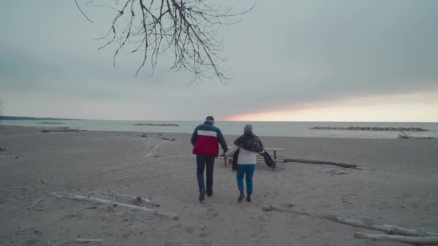 A couple wearing jackets walks on the beach toward the ocean on a cloudy day