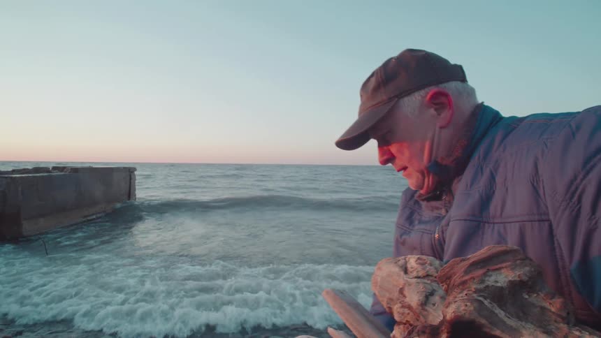 An elderly man picks up rubble from the beach during the day as waves hit the shore