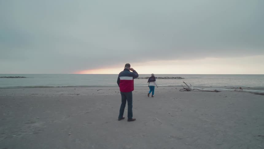 Two people walks on a beach near the water at dusk one taking a photo of the sunset