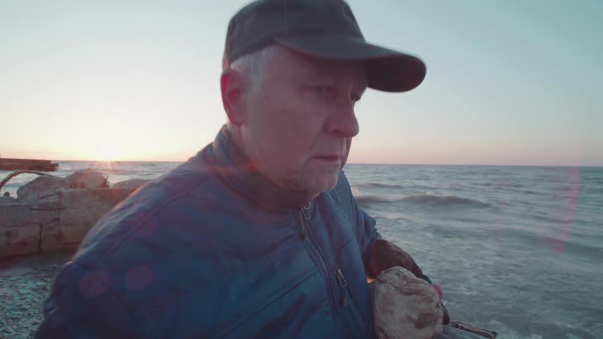A man walks along a rocky beach at sunset holding firewood for a campfire