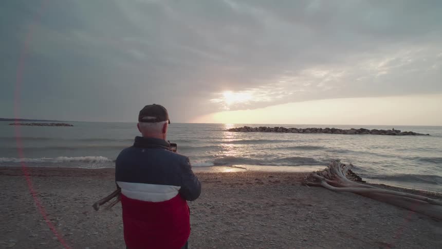 A man stands on the beach taking a photo of the sunset over the ocean