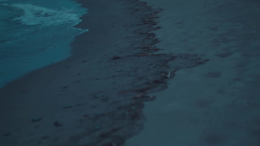 Waves gently wash upon a dark sand beach under a dim ambient light