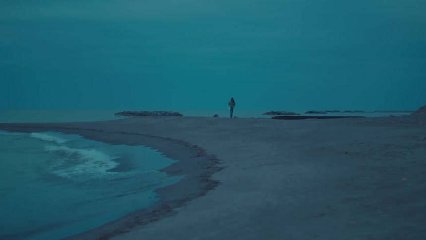 Woman wearing a hooded jacket and carrying a bag walks on the beach as waves roll in to shore