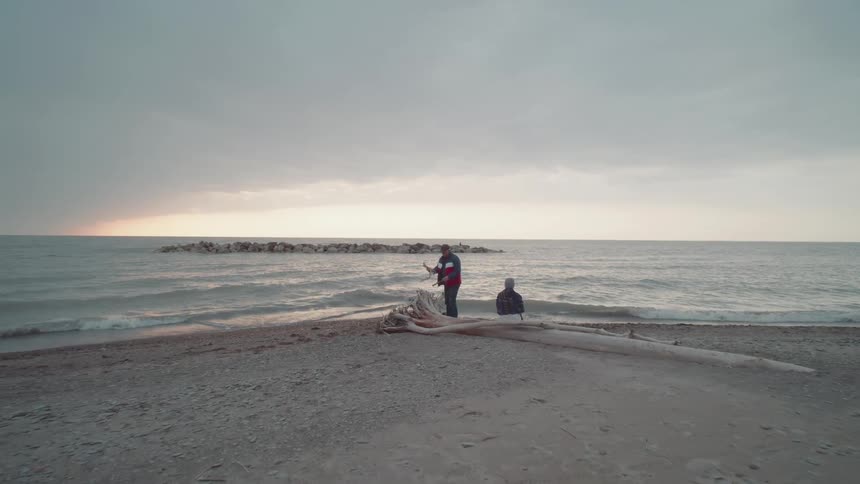 Two people relax by driftwood at a rocky beach during sunset
