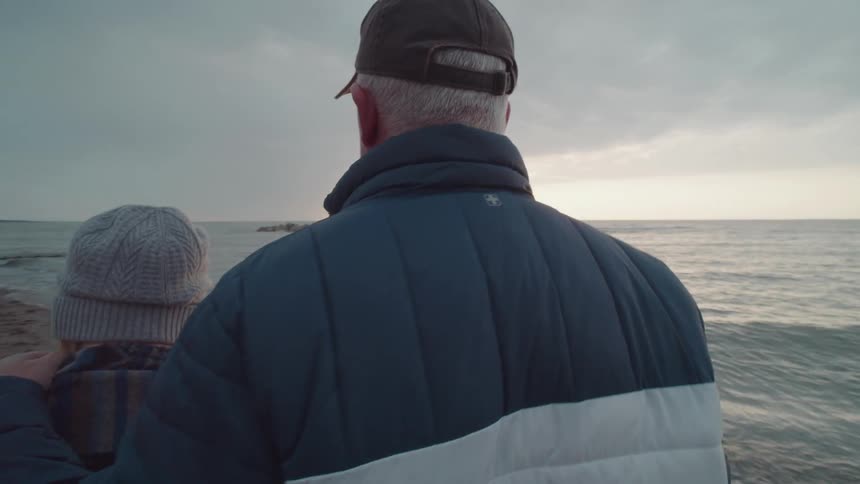Two people wearing winter clothing stand close together facing the ocean at sunset