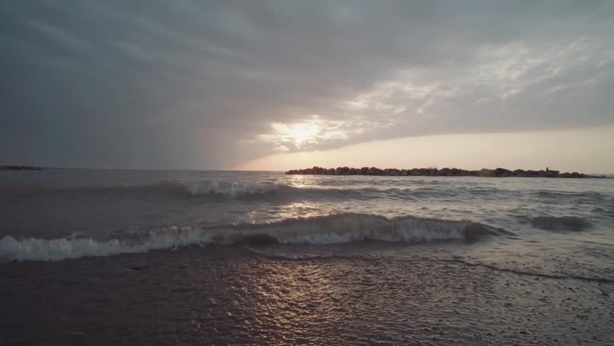 Under a dramatic overcast sky gentle waves roll onto a dark wet shore illuminated by the suns reflection on the water