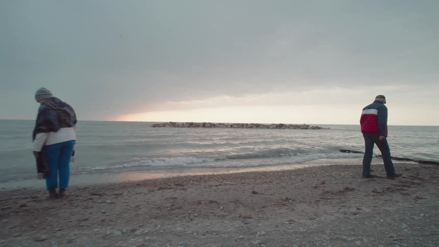 A couple both wearing jackets walks along the shore as ocean waves hit the beach on a cloudy day