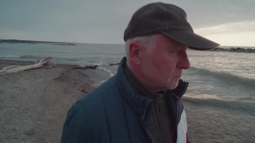 An older man in a cap gazes out over the water at sunset from a quiet beach