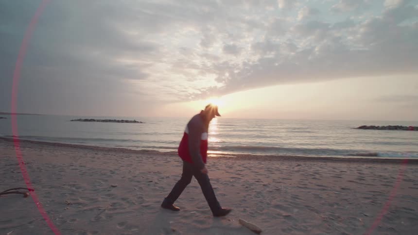 Man walking on the beach picks up a piece of driftwood as waves roll in to shore and the sun glows on the horizon