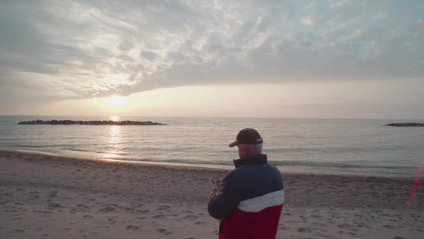 An elderly man walks on the beach sand at sunset pulls out his smartphone and starts recording the ocean view while wearing a jacket and a baseball hat