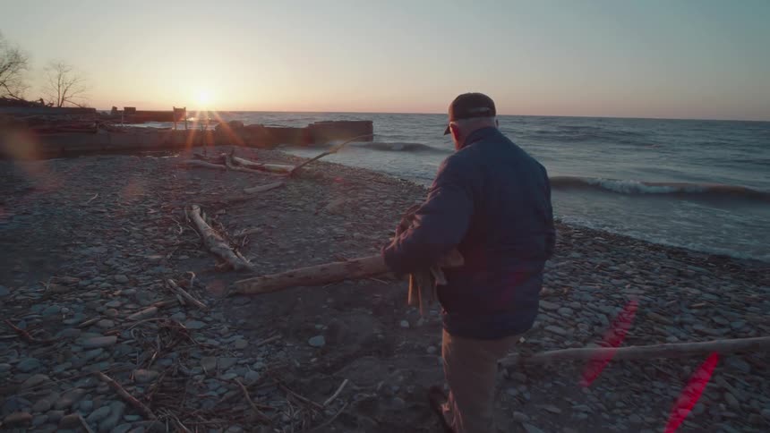 An older man collects firewood along a rocky shoreline at sunset