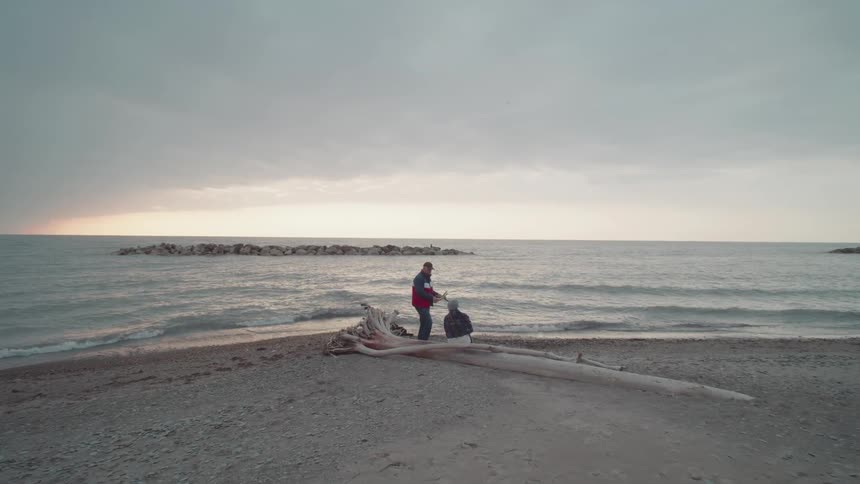 Two people quietly fish by a calm shoreline under a cloudy sky