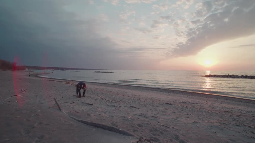 A lone person walks along a sandy driftwood strewn beach as the sun sets over the ocean