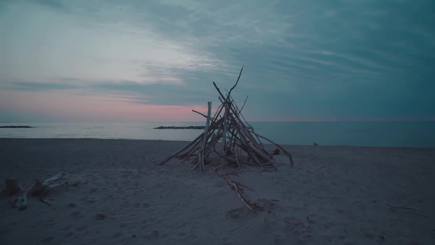 A pile of driftwood stands on an empty beach at dusk under a soft blue and pink sky