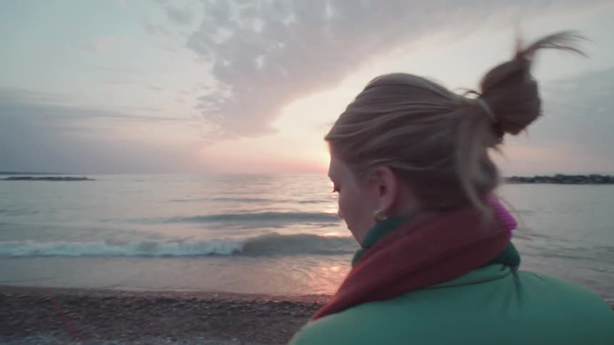 A woman stands on a quiet beach at sunset gazing out toward the horizon