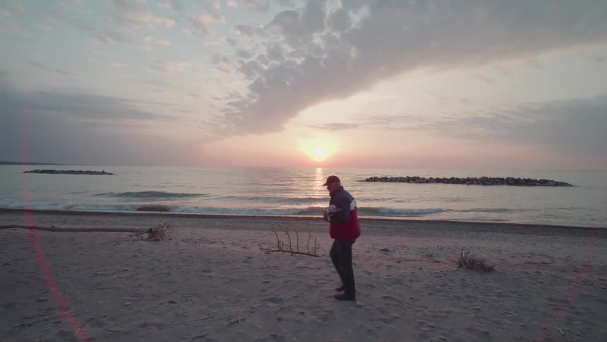 A man walks along a quiet beach at sunrise facing the gentle waves and pastel sky