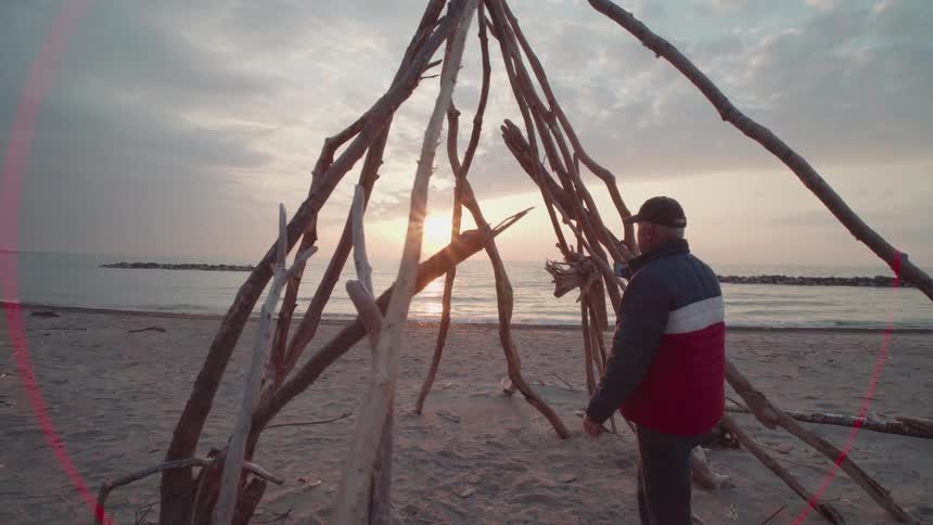 Man takes a picture of the sunset at the beach through the bones of a tepee