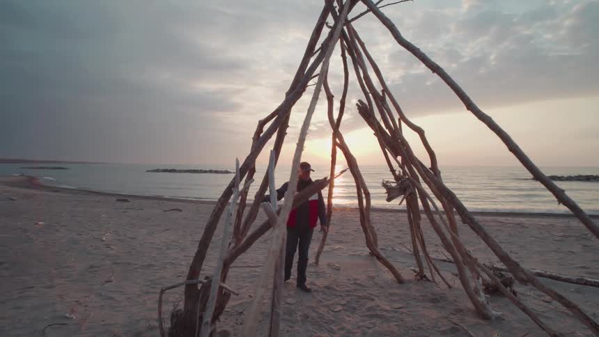 Man walks on the beach to a structure built from driftwood