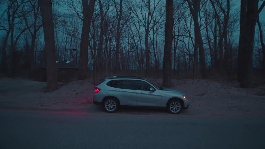 A silver suv is parked beside a quiet road in a forested area at dusk