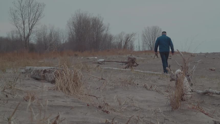 A man walks away through a desolate sandy landscape scattered with driftwood on an overcast day