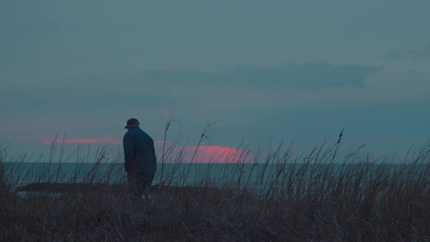 Silhouetted at dusk a man in a jacket walks along a grassy beach and stops to take a picture of the sunset over the ocean