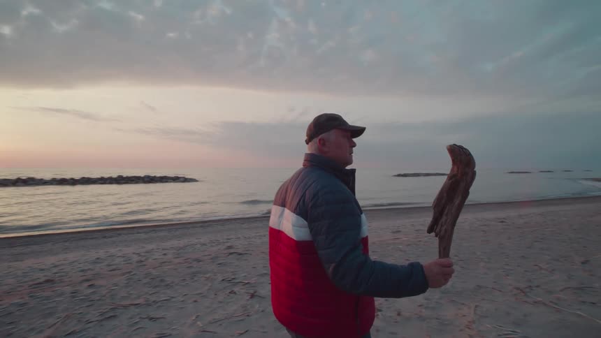 Man walks along the beach holding a piece of wood at dusk