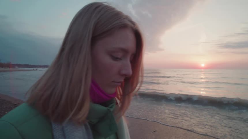 A young woman walks pensively along the beach at sunset