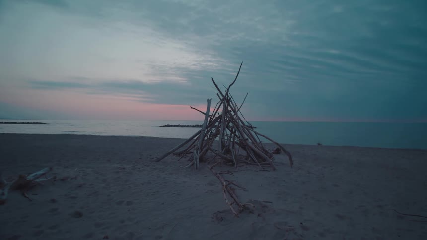 A driftwood teepee structure stands on a quiet beach under a dusky sky