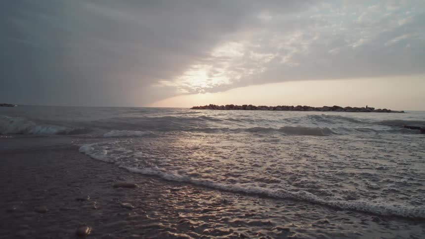 Gentle waves wash over a pebble beach as the sun begins to break through an overcast sky creating a golden reflection on the waters surface