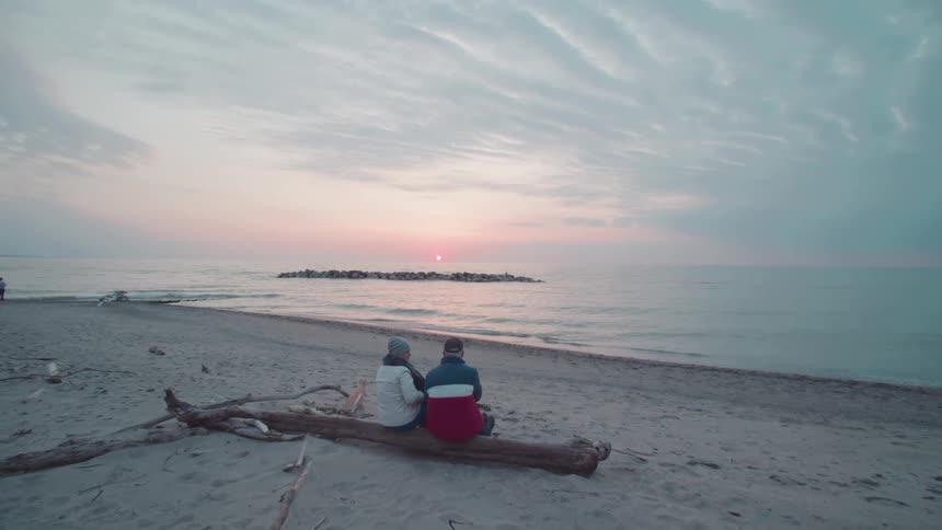 An elderly couple both wearing jackets sits on a log of wood watching the sea as waves hit the shore around midday