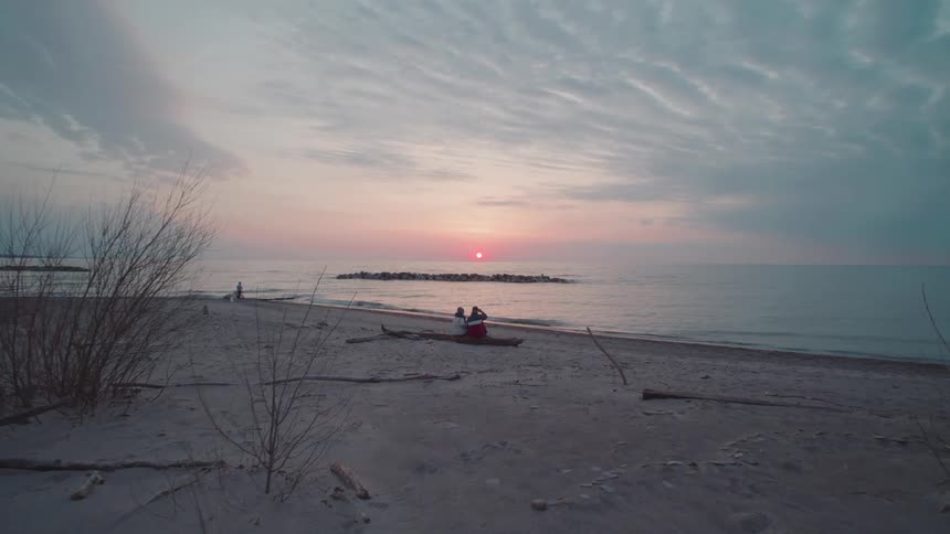 Two people sitting on the beach watching waves roll in to shore as the sun glows on the horizon
