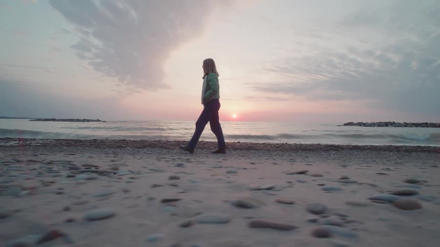 Woman walks across the beach as waves roll in to shore and the sun glows on the horizon