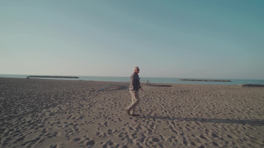 An older man walks alone across a sandy beach under a clear blue sky