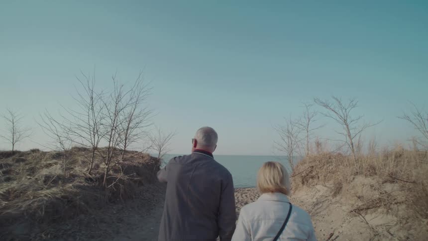 Back of elderly man and woman walking towards the beach at midday with their hands held together