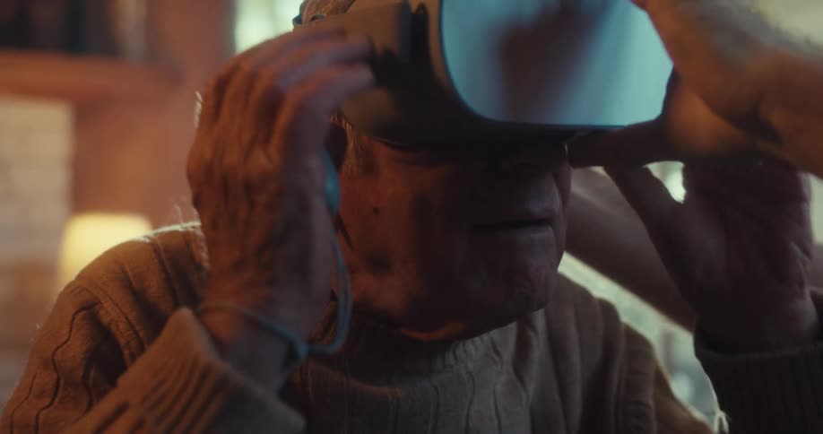 An elderly man is helped by another person to wear a vr headset indoors around midday