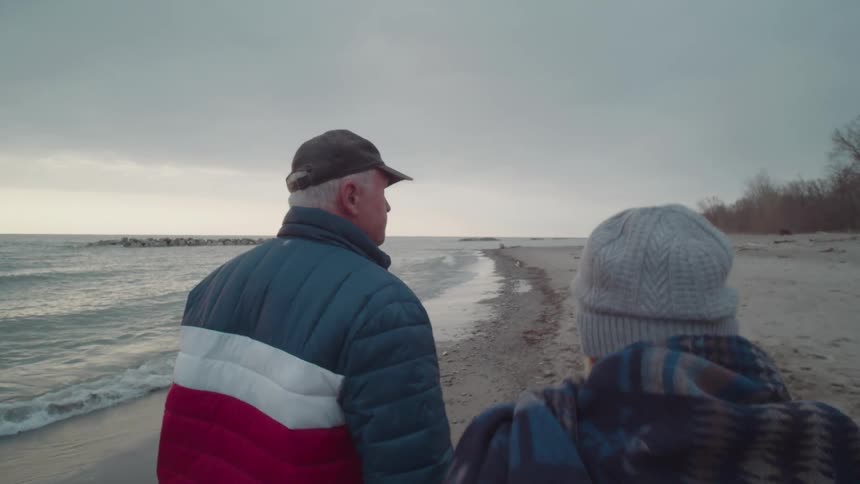 An older couple bundled up in jackets and hats walks along a quiet sandy beach on a cool overcast day