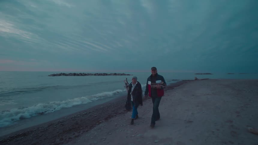 An elderly couple walks side by side on the beach at sundown each carrying sticks for firewood and wearing jackets and hats as ocean waves hit the shore