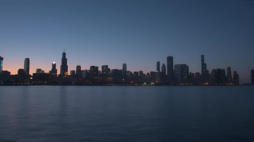 The sun sets behind a city skyline as small waves ripple through the water