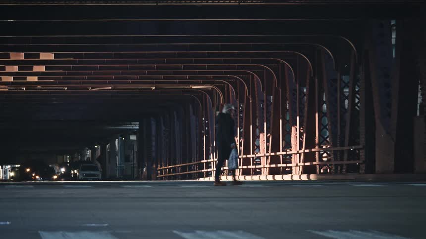 A person carrying a bag walks across a crosswalk on a street running under a large industrial city bridge at dusk