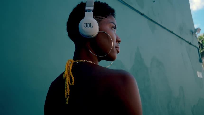 A woman poses to camera wearing headphones modeling against an urban wall backdrop