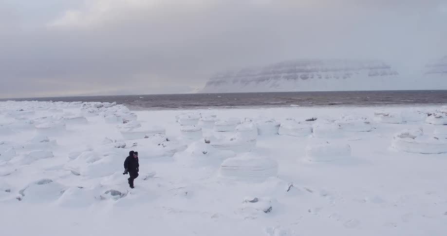 A man carrying a camera walks along the icy snow covered shore