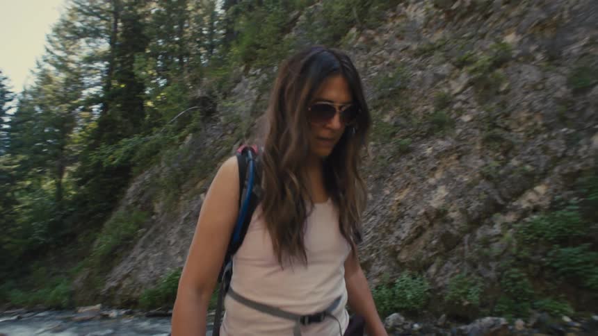 A woman hikes through a shallow stream in a forested mountain area