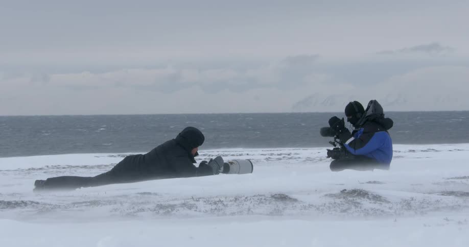Two people with cameras sit in the snow by a body of water with a cloudy sky